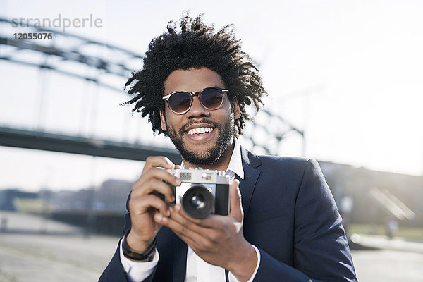 Lächelnder Mann im Anzug am Flussufer mit einer Vintage-Kamera