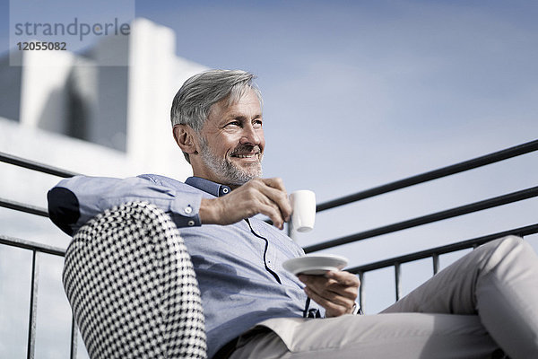 Lächelnder grauhaariger Mann sitzt auf dem Balkon und trinkt Kaffee.
