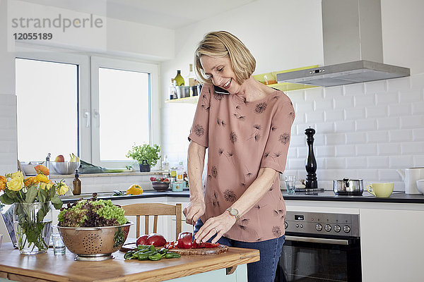Reife Frau bereitet einen Salat zu und telefoniert in der Küche.
