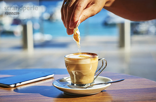 Nahaufnahme der Frauenhand  die Zucker in ein Glas Espresso Macchiato gießt.