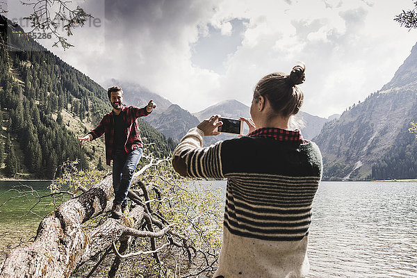 Österreich  Tirol  Alpen  Frau macht Handyfoto von Mann beim Balancieren auf Baumstamm am Bergsee