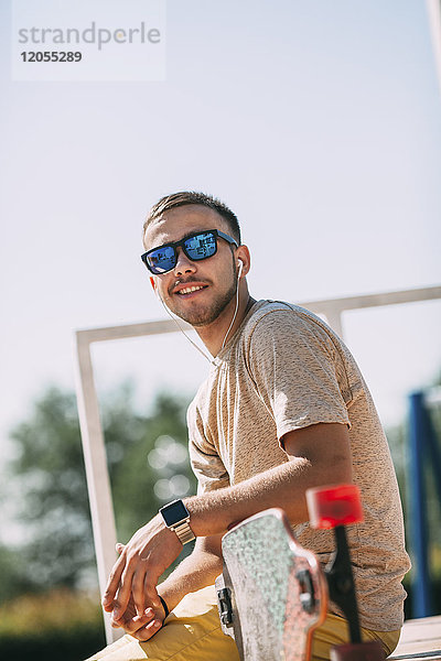 Lächelnder junger Mann mit Ohrstöpseln und Longboard im Skatepark