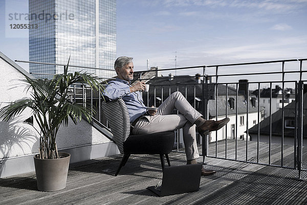 Grauhaariger Geschäftsmann auf dem Balkon mit Blick über die Stadt und Kaffeetrinken