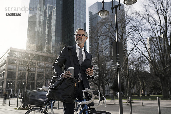 Grauhaariger Geschäftsmann mit Fahrrad und Kaffee zum Mitnehmen in der Stadt