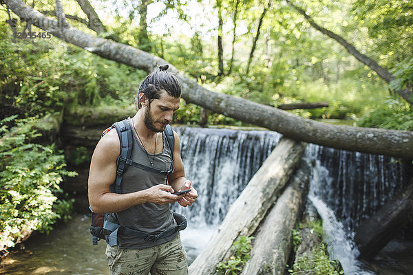 Junger Mann mit Handy an einem Wasserfall im Wald