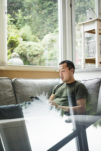 Mann zu Hause mit Tablette auf der Couch neben dem Fenster