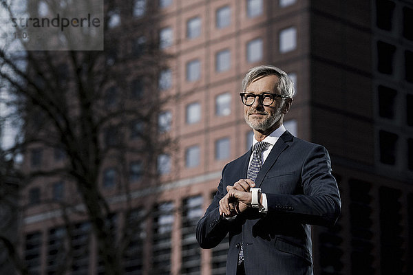 Grauhaariger Geschäftsmann vor rotem Wolkenkratzer  der auf seine Smartwatch zeigt.