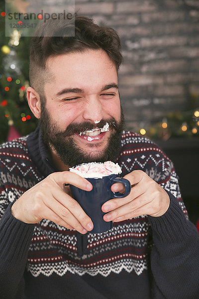 Porträt eines lachenden Mannes  der heiße Schokolade mit Schlagsahne und gehackten Zuckerstangen trinkt.