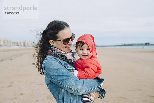 Porträt des lächelnden Mädchens auf den Armen ihrer Mutter am Strand