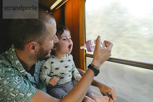 Vater gibt seinem kleinen Mädchen Wasser in einer Babyflasche  während es mit dem Zug unterwegs ist.