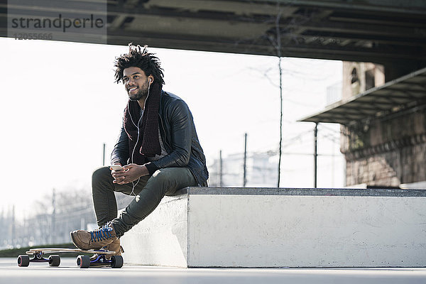 Lächelnder Mann mit Longboard sitzt im Skatepark unter der Brücke und hält ein Smartphone.