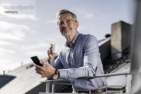 Grauhaariger Geschäftsmann mit Smartphone auf dem Balkon beim Kaffeetrinken