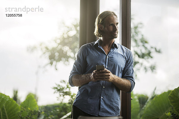 Hübscher Mann mit Smartphone in einem modernen Designhaus mit Glasfassade  umgeben von einem üppigen tropischen Garten.