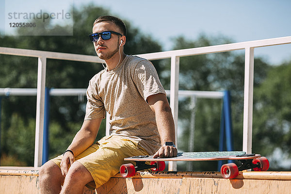 Junger Mann mit Ohrstöpseln und Longboard im Skatepark sitzend