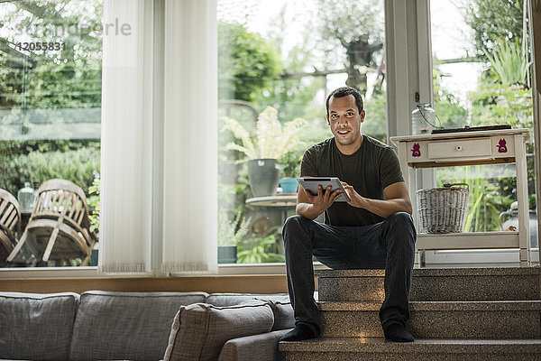Porträt eines lächelnden Mannes zu Hause  der auf einer Treppe mit Tablette sitzt.