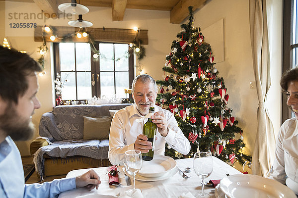 Lächelnder älterer Mann mit Familie hält Flasche Wein am Weihnachtstisch