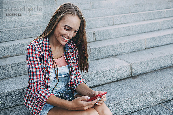 Lächelnde junge Frau sitzt auf der Treppe und schaut aufs Handy.