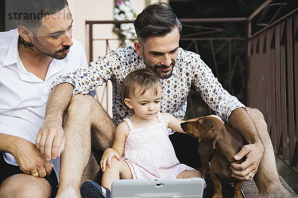 Schwules Paar mit Tochter und Hund auf dem Balkon mit digitalem Tablett