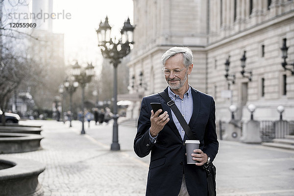 Grauhaariger Geschäftsmann in der Stadt mit Smartphone und Kaffee zum Mitnehmen