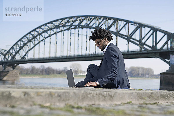 Geschäftsmann sitzend am Flussufer mit Laptop