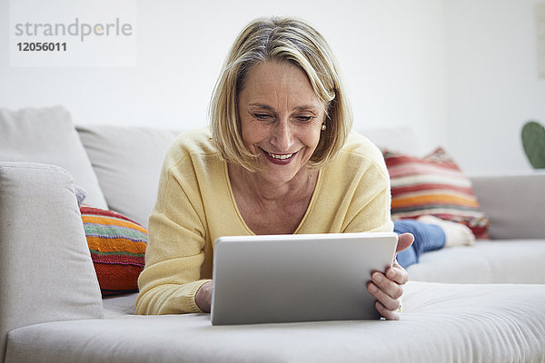 Lächelnde reife Frau zu Hause mit Tablette auf dem Sofa