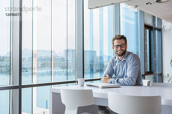 Porträt eines lächelnden Mannes mit Laptop und Tasse Kaffee im modernen Büro