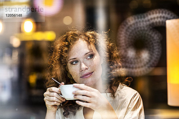 Porträt einer lächelnden jungen Frau mit Kaffeetasse hinter der Fensterscheibe eines Cafés