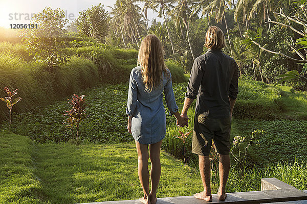 Ein Paar hält sich an den Händen  während es am Rande eines Pools steht und einen atemberaubenden Blick auf den Sonnenuntergang in einem üppigen tropischen Garten genießt.