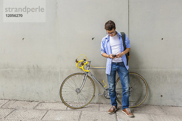Junger Mann mit Rennrad beim Blick auf das Handy vor der Betonwand