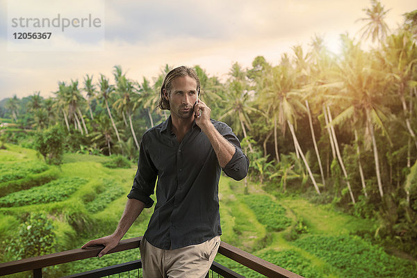 Gutaussehender Mann  der sich auf den Balkon stützt und auf dem Smartphone spricht mit atemberaubender Aussicht auf die tropische Landschaft.