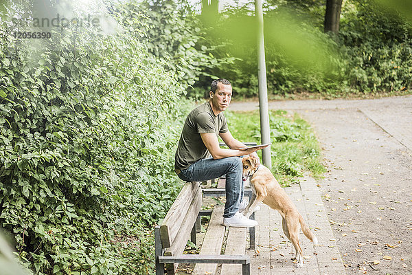 Porträt eines ernsthaften Mannes mit Tablette und Hund auf einer Bank im Park