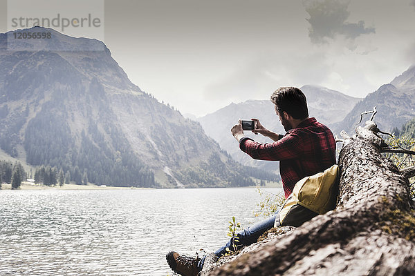 Österreich  Tirol  Alpen  Wanderer auf Baumstamm am Bergsee sitzend  Handyfoto machend