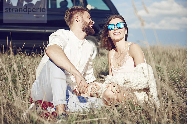 Glückliches junges Paar sitzt auf dem Feld neben dem Auto