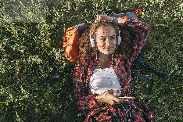 Teenagermädchen mit Rucksack auf einer Wiese liegend Musik hören mit Kopfhörer und Handy