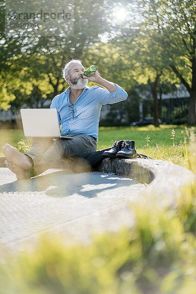 Erwachsener Mann trinkt Bier und benutzt Laptop im Park