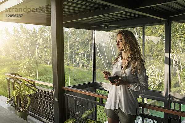 Lächelnde Frau isst Müsli in einem modernen Designhaus mit Pool und Glasfassade  umgeben von einem üppigen tropischen Garten.