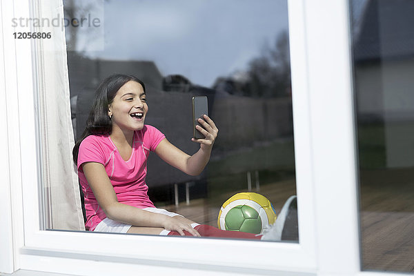 Fröhliches Mädchen im Fußball-Outfit sitzt auf dem Boden im Wohnzimmer und nimmt einen Selfie