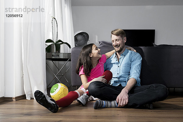 Mädchen im Fußball-Outfit neben Vater auf dem Boden im Wohnzimmer sitzend