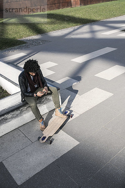 Mann sitzend im Skatepark mit Longboard Tippen auf dem Smartphone
