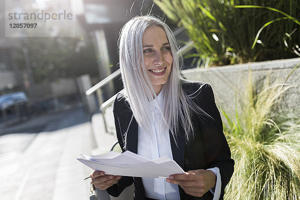 Lächelnde junge Geschäftsfrau auf einer Mauer in der Stadt sitzend mit Dokumenten