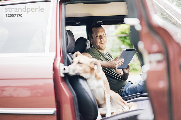 Porträt des lächelnden Mannes mit Tablette im Auto mit Hund auf Beifahrersitz