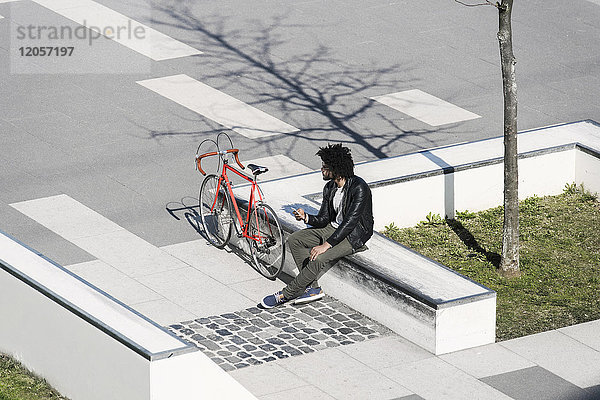 Mann sitzt im Skatepark und hält sein Smartphone neben dem Fahrrad.
