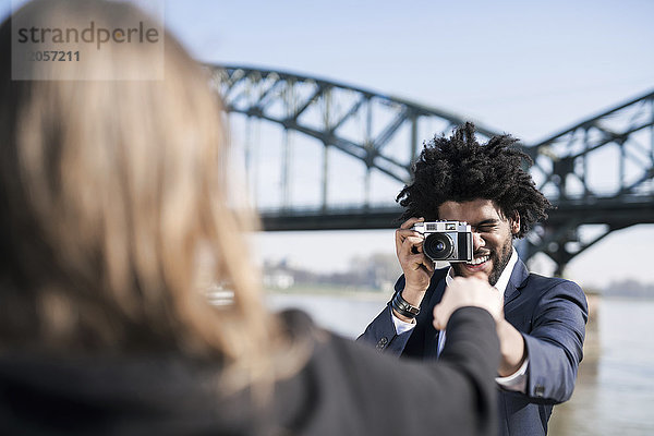 Lächelnder Mann im Anzug am Flussufer beim Fotografieren seiner Freundin mit einer alten Kamera
