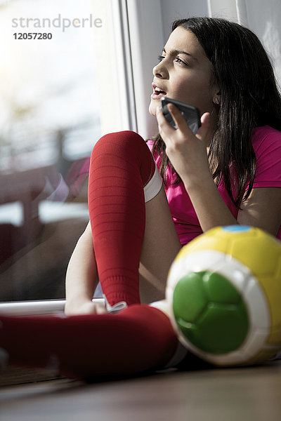 Mädchen im Fußball-Outfit sitzt auf dem Boden im Wohnzimmer und spricht mit jemandem auf ihrem Smartphone.