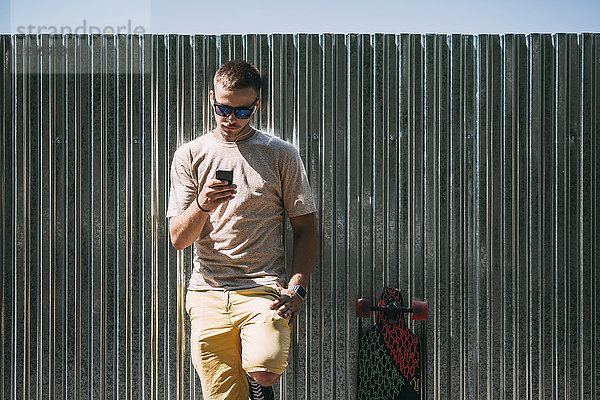 Junger Mann mit Handy und Ohrstöpseln  die an einer Wand neben dem Longboard lehnen