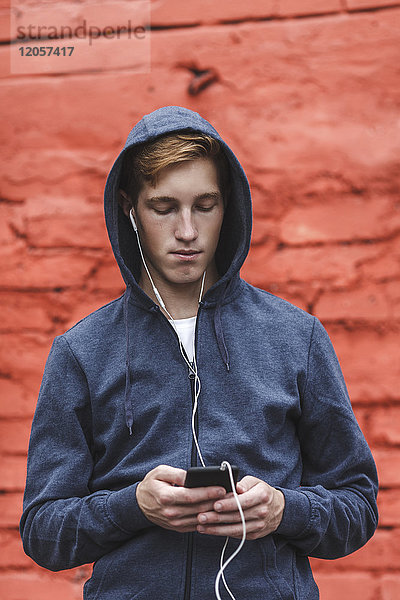 Junger Mann mit Handy und Kopfhörer an roter Ziegelwand