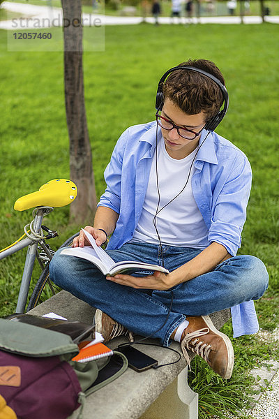 Junger Mann mit Rennrad und Kopfhörer auf einer Bank sitzend  Buch lesend