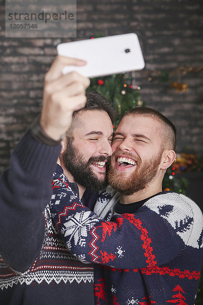 Porträt eines lachenden schwulen Paares  das sich zu Hause mit dem Smartphone in die Weihnachtszeit begibt.