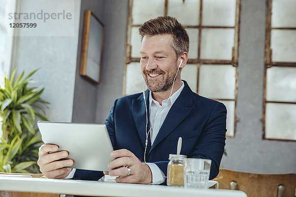 Lächelnder Geschäftsmann mit Tablette und Ohrstöpseln im Cafe