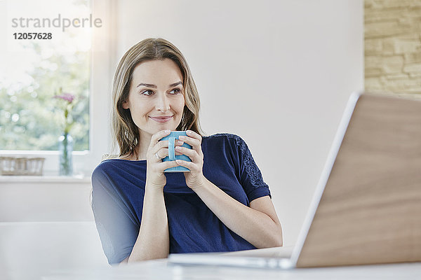 Lächelnde Frau zu Hause mit Kaffeetasse und Laptop